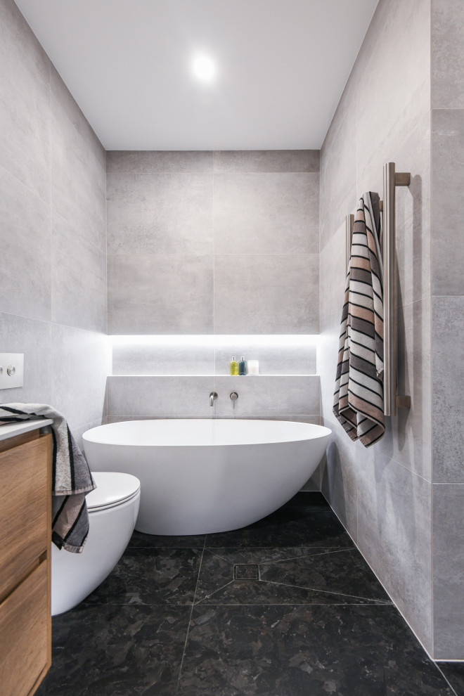 Idéer för mellanstora funkis vitt en-suite badrum, med släta luckor, skåp i mellenmörkt trä, ett fristående badkar, en öppen dusch, en vägghängd toalettstol, svart kakel, grå kakel, keramikplattor, grå väggar, klinkergolv i keramik, ett nedsänkt handfat, bänkskiva i akrylsten, svart golv och med dusch som är öppen