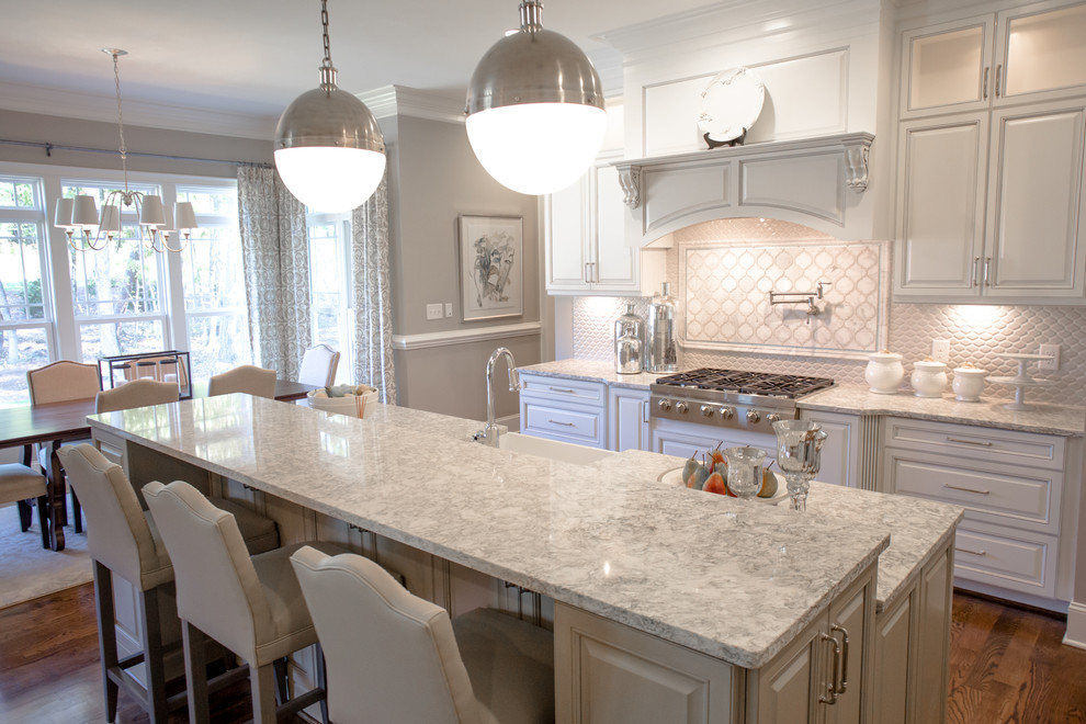 This is an example of a transitional l-shaped open plan kitchen in Raleigh with a farmhouse sink, raised-panel cabinets, white cabinets, quartzite benchtops, white splashback, ceramic splashback, stainless steel appliances, medium hardwood floors and with island.