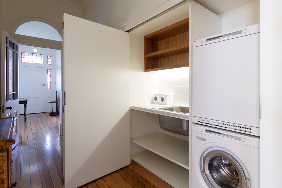 This is an example of a small traditional single-wall utility room in Hobart with a drop-in sink, open cabinets, white cabinets, marble benchtops, white walls, light hardwood floors and a stacked washer and dryer.