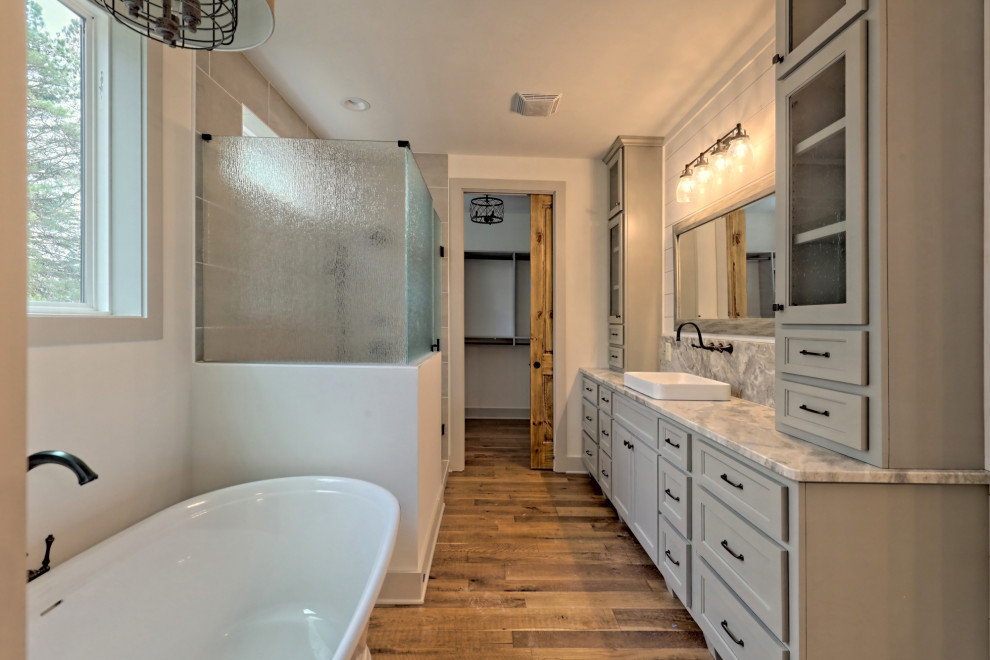 Photo of a large country master bathroom in Atlanta with recessed-panel cabinets, grey cabinets, a freestanding tub, an alcove shower, a two-piece toilet, white walls, medium hardwood floors, a vessel sink, granite benchtops, brown floor, a hinged shower door, grey benchtops, an enclosed toilet, a double vanity, a built-in vanity and wood walls.