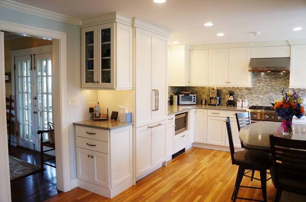 Mid-sized transitional l-shaped eat-in kitchen in Boston with a farmhouse sink, shaker cabinets, white cabinets, quartz benchtops, blue splashback, glass tile splashback, panelled appliances, light hardwood floors and a peninsula.
