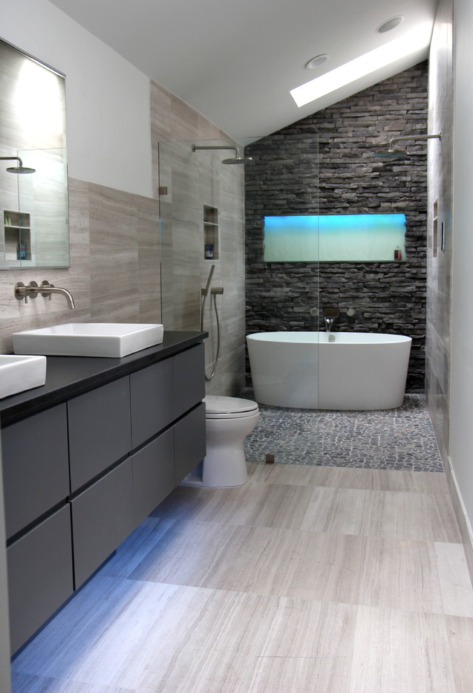 Photo of a mid-sized contemporary master bathroom in Atlanta with a vessel sink, grey cabinets, a freestanding tub, a curbless shower, a two-piece toilet, gray tile, flat-panel cabinets, stone tile, white walls, limestone floors, granite benchtops, grey floor, an open shower, black benchtops, a niche, a double vanity, a floating vanity and vaulted.