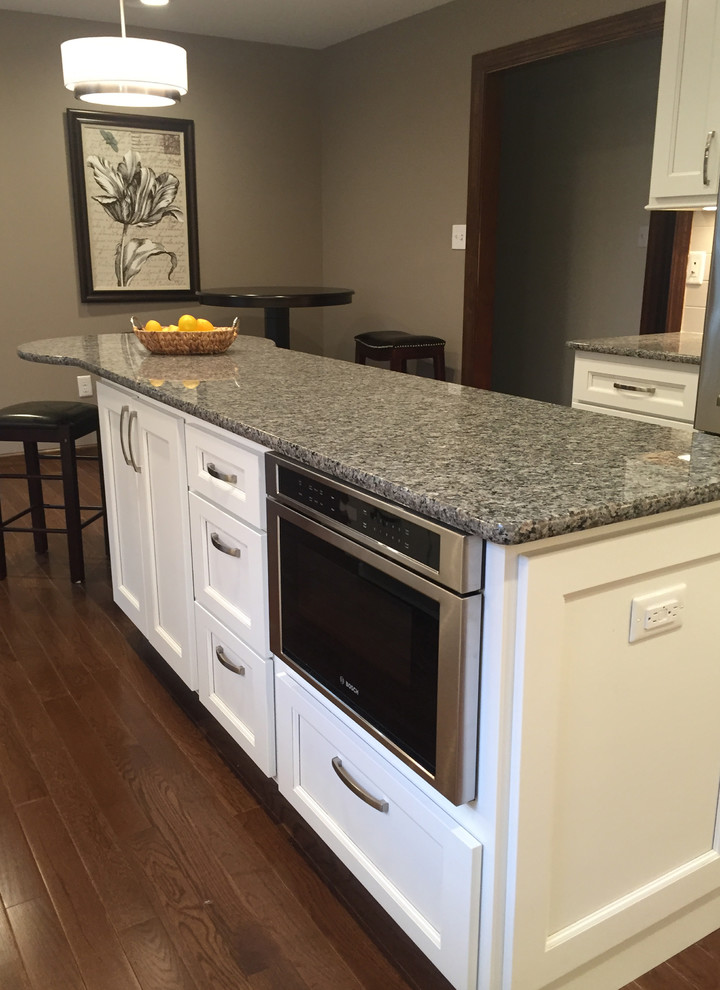 Example of a transitional galley dark wood floor kitchen design with an undermount sink, flat-panel cabinets, white cabinets, granite countertops, white backsplash, porcelain backsplash, stainless steel appliances and an island
