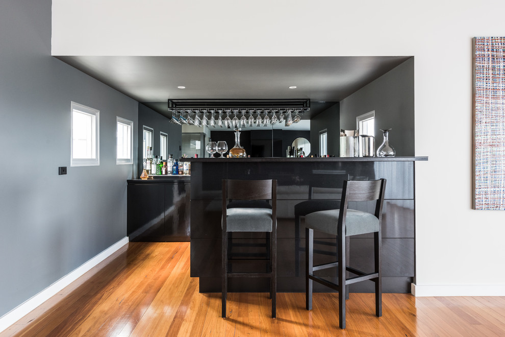 Photo of a contemporary galley wet bar in Melbourne with flat-panel cabinets, black cabinets, mirror splashback, medium hardwood floors, brown floor and black benchtop.