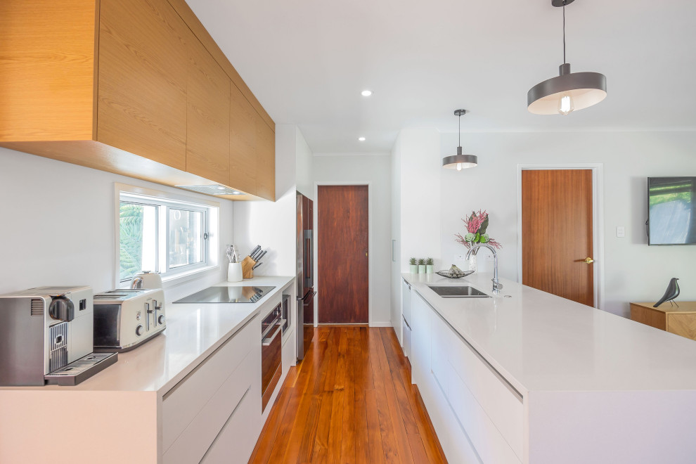 Zweizeilige, Kleine Eklektische Wohnküche mit Unterbauwaschbecken, flächenbündigen Schrankfronten, weißen Schränken, Mineralwerkstoff-Arbeitsplatte, Küchenrückwand in Weiß, braunem Holzboden, Halbinsel, braunem Boden und weißer Arbeitsplatte in Auckland
