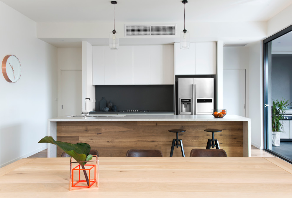 Photo of a large contemporary galley open plan kitchen in Melbourne with a double-bowl sink, flat-panel cabinets, white cabinets, quartz benchtops, black splashback, stone slab splashback, stainless steel appliances, light hardwood floors and with island.