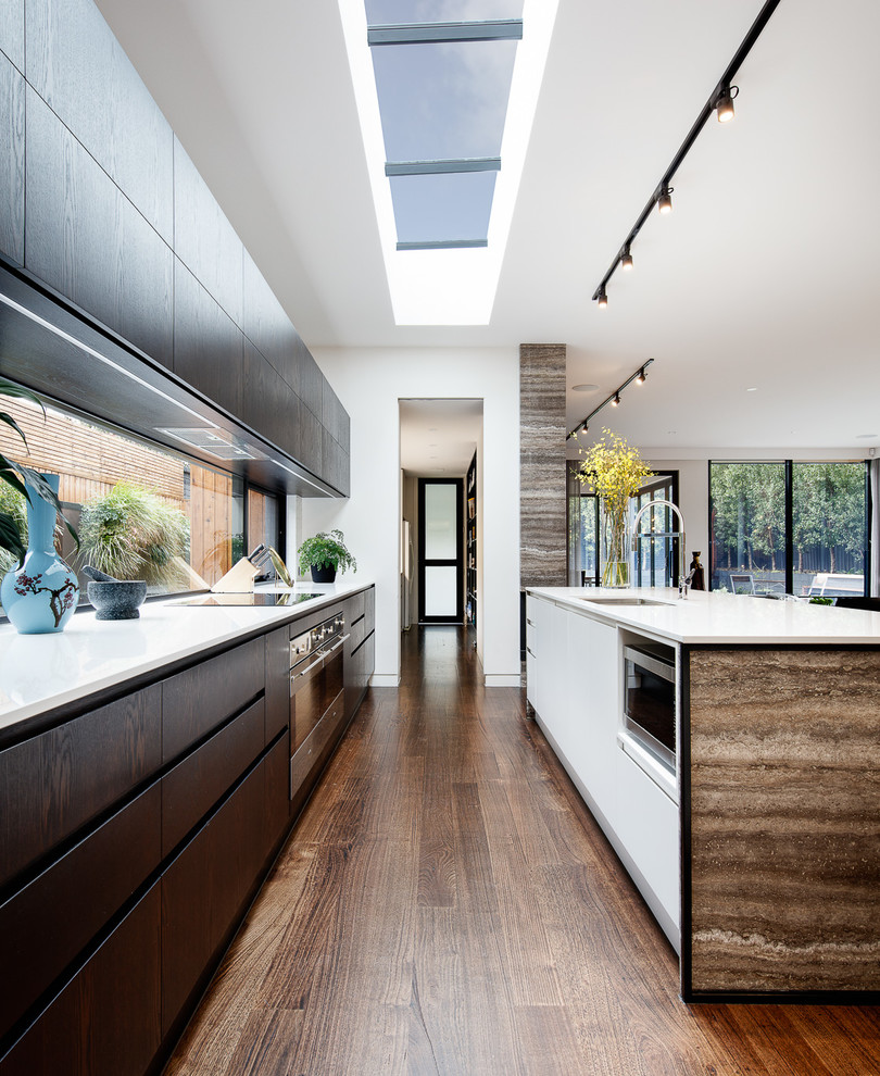 This is an example of a large contemporary galley kitchen pantry in Melbourne with a double-bowl sink, flat-panel cabinets, dark wood cabinets, glass sheet splashback, stainless steel appliances, dark hardwood floors, with island, brown floor and white benchtop.