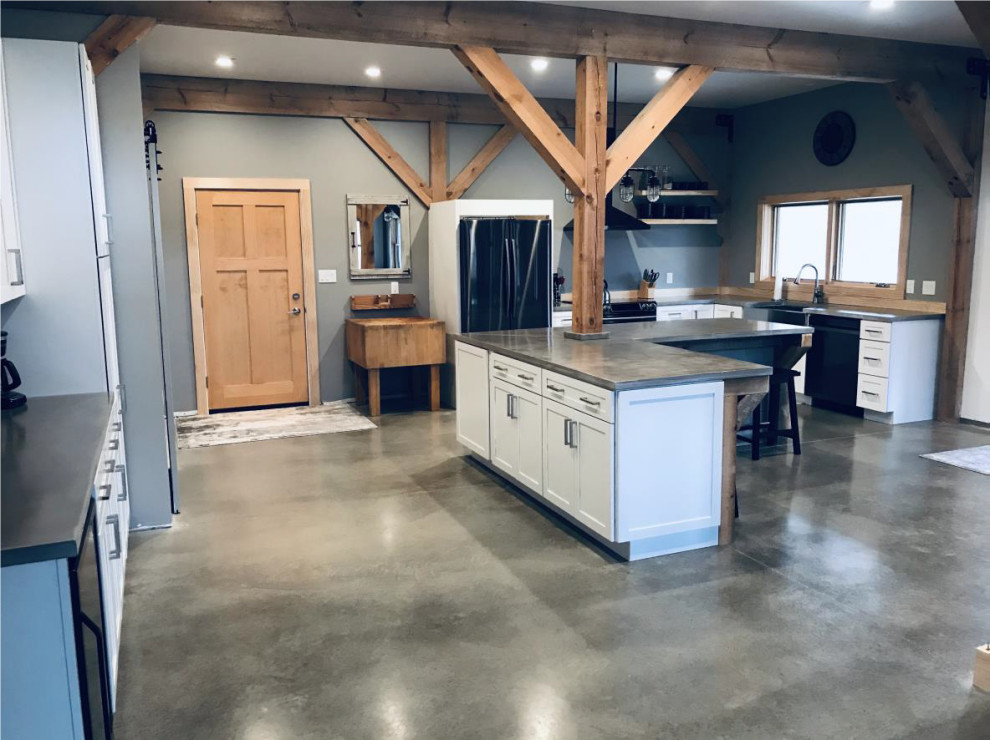 Photo of a large country l-shaped kitchen/diner in Chicago with a belfast sink, shaker cabinets, white cabinets, concrete worktops, black appliances, concrete flooring, an island, grey floors, grey worktops and exposed beams.