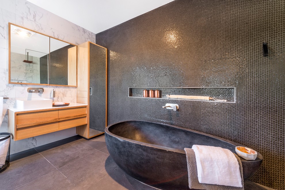 Design ideas for a large contemporary master bathroom in Melbourne with recessed-panel cabinets, light wood cabinets, a freestanding tub, a double shower, a wall-mount toilet, gray tile, glass tile, white walls, ceramic floors, a vessel sink and solid surface benchtops.