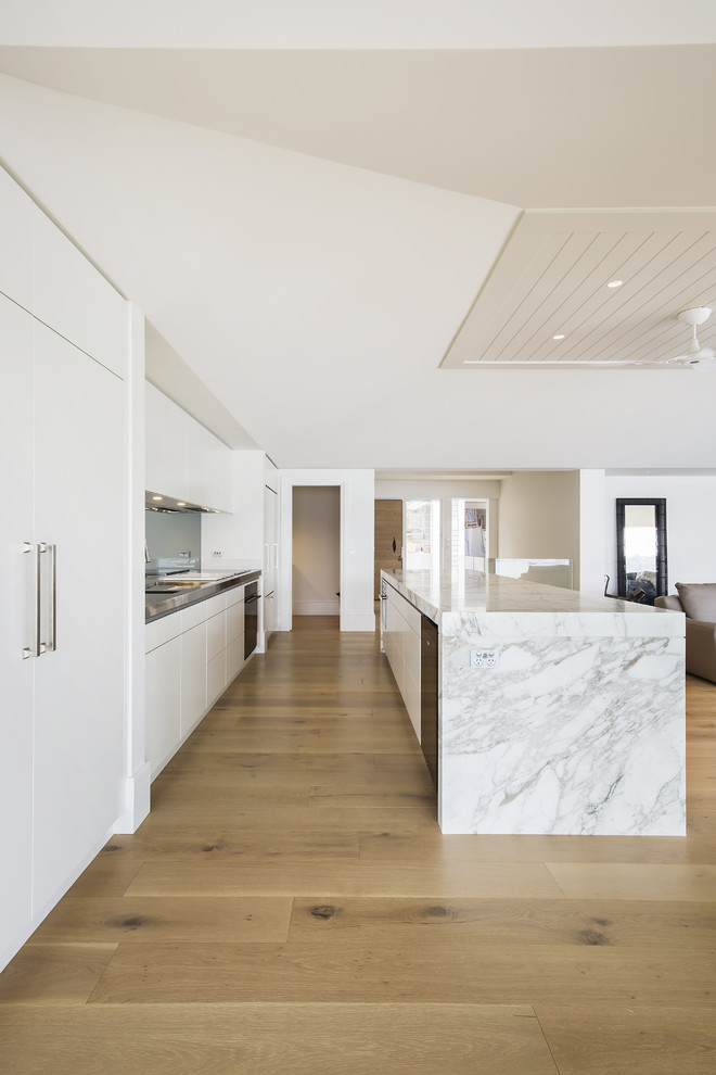 Inspiration for a large modern single-wall open plan kitchen in Sydney with an integrated sink, flat-panel cabinets, beige cabinets, marble benchtops, metallic splashback, mirror splashback, black appliances, light hardwood floors and with island.