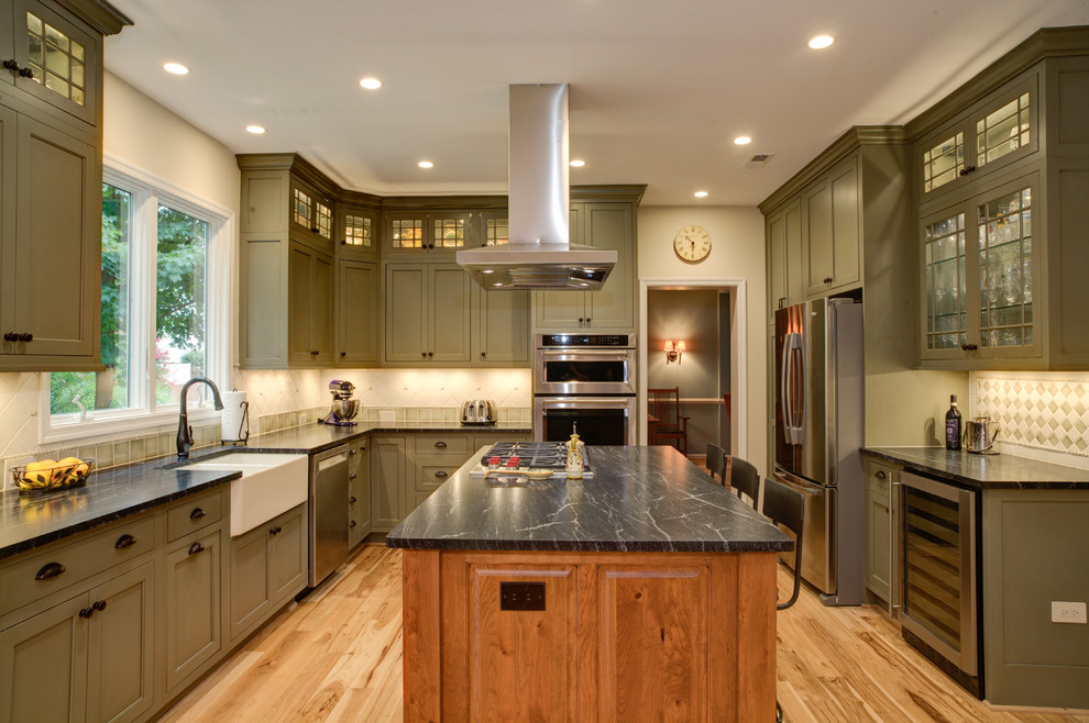 Photo of a large country u-shaped separate kitchen in DC Metro with a farmhouse sink, shaker cabinets, green cabinets, soapstone benchtops, beige splashback, ceramic splashback, stainless steel appliances, light hardwood floors and with island.