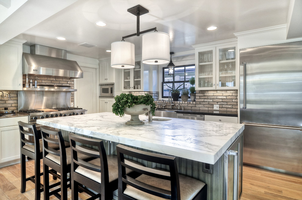 Design ideas for a mid-sized transitional l-shaped open plan kitchen in Orange County with with island, a farmhouse sink, glass-front cabinets, white cabinets, marble benchtops, beige splashback, brick splashback, stainless steel appliances and light hardwood floors.