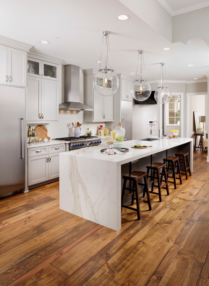Mid-sized transitional galley open plan kitchen in Houston with a farmhouse sink, shaker cabinets, grey cabinets, white splashback, stainless steel appliances, medium hardwood floors, with island, quartz benchtops, brown floor and white benchtop.