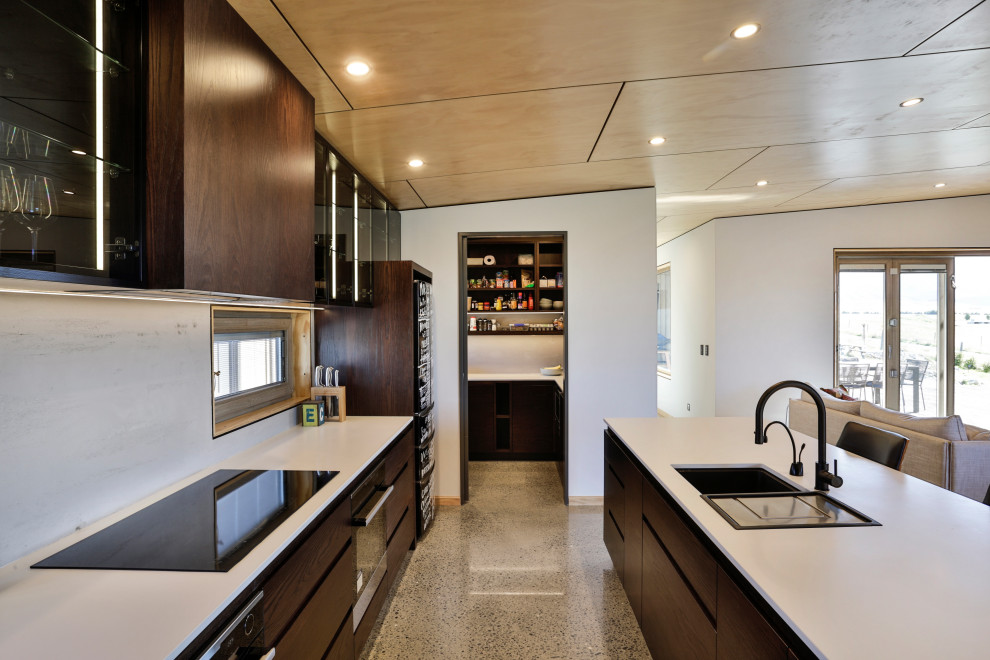 Design ideas for a mid-sized contemporary single-wall open plan kitchen in Christchurch with an undermount sink, flat-panel cabinets, dark wood cabinets, quartz benchtops, grey splashback, black appliances, concrete floors, with island, grey floor and white benchtop.
