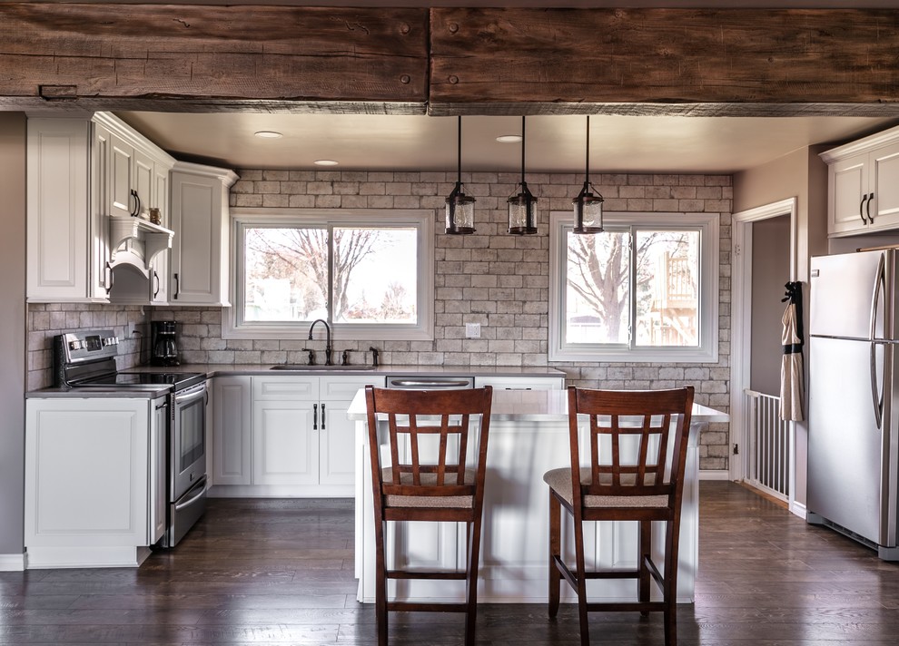 Mid-sized mountain style u-shaped laminate floor and brown floor open concept kitchen photo with an undermount sink, raised-panel cabinets, white cabinets, quartzite countertops, white backsplash, brick backsplash, stainless steel appliances, an island and gray countertops