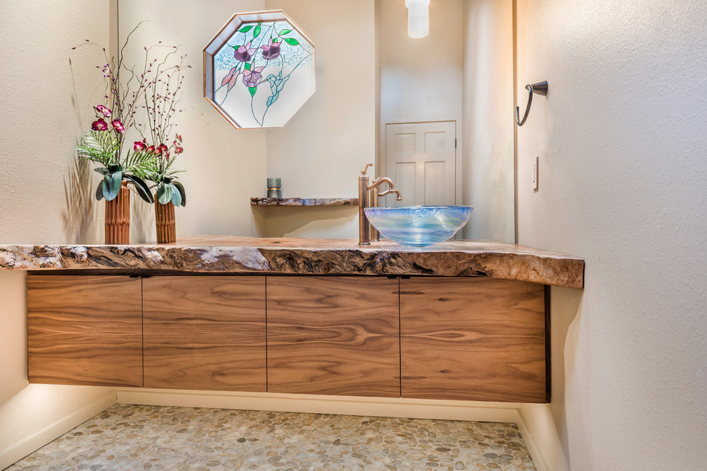 This is an example of a mid-sized country powder room in San Diego with flat-panel cabinets, medium wood cabinets, multi-coloured tile, beige walls, pebble tile floors, a vessel sink and wood benchtops.