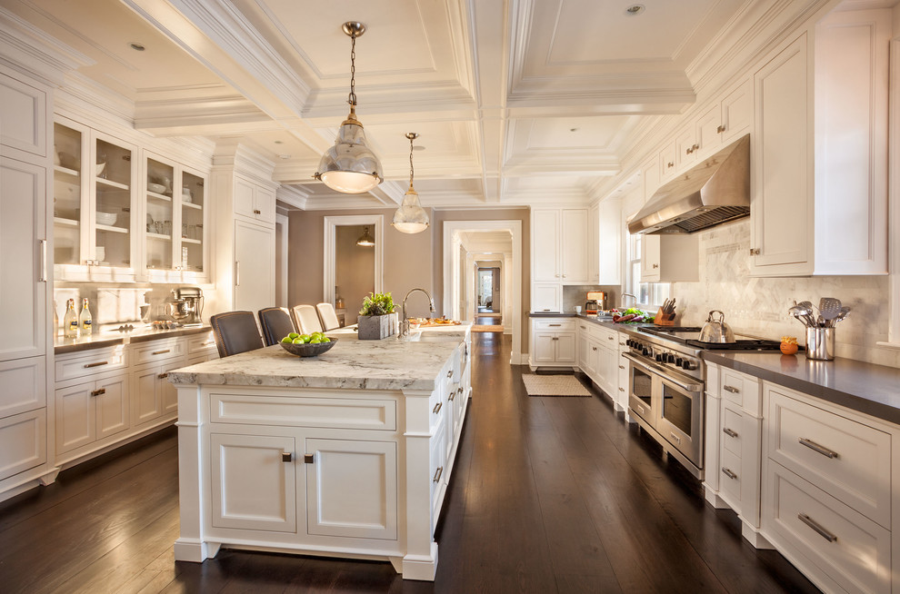 Large traditional galley separate kitchen in New York with a farmhouse sink, white cabinets, marble benchtops, white splashback, stone tile splashback, stainless steel appliances, dark hardwood floors, with island and recessed-panel cabinets.