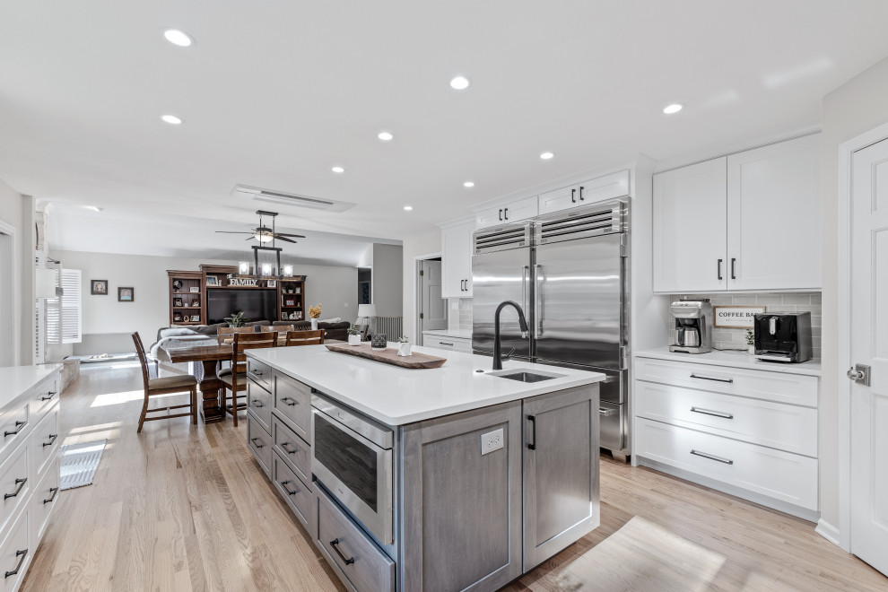 Inspiration for a large transitional u-shaped light wood floor and multicolored floor eat-in kitchen remodel with a single-bowl sink, shaker cabinets, white cabinets, quartz countertops, white backsplash, quartz backsplash, stainless steel appliances, an island and white countertops