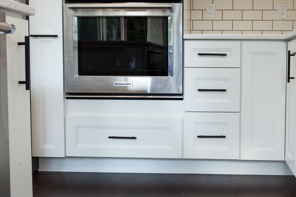 Home design - large transitional l-shaped dark wood floor and brown floor home design idea with an undermount sink, recessed-panel cabinets, white cabinets, quartz countertops, white backsplash, subway tile backsplash, stainless steel appliances and an island