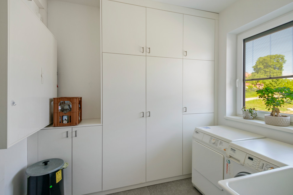 Inspiration for a mid-sized contemporary l-shaped utility room in Hamburg with a single-bowl sink, flat-panel cabinets, white cabinets, white walls, vinyl floors, a side-by-side washer and dryer, grey floor and white benchtop.