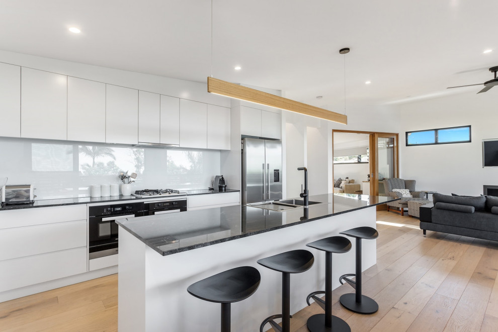 Contemporary open plan kitchen in Sydney with a double-bowl sink, flat-panel cabinets, white cabinets, white splashback, glass sheet splashback, stainless steel appliances, light hardwood floors, with island and black benchtop.