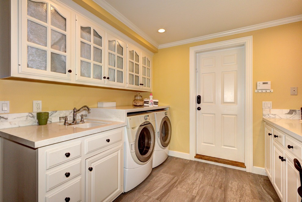 Large traditional galley dedicated laundry room in San Francisco with an undermount sink, raised-panel cabinets, white cabinets, solid surface benchtops, yellow walls, porcelain floors, a side-by-side washer and dryer, brown floor and white benchtop.