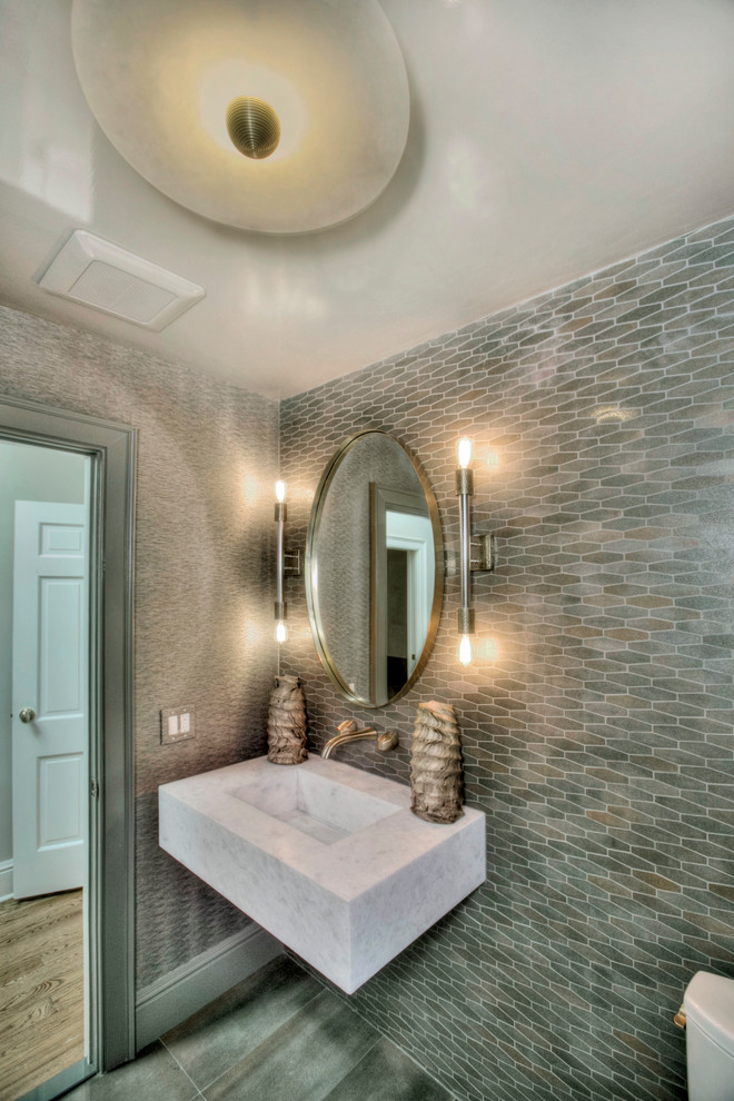 Mid-sized transitional powder room in New York with a one-piece toilet, gray tile, porcelain tile, grey walls, porcelain floors, a wall-mount sink, marble benchtops, grey floor and white benchtops.