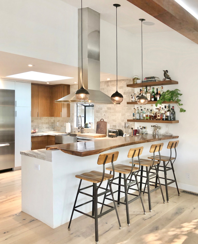 This is an example of a contemporary u-shaped open plan kitchen in San Francisco with a farmhouse sink, flat-panel cabinets, medium wood cabinets, marble benchtops, grey splashback, subway tile splashback, stainless steel appliances, light hardwood floors, a peninsula, beige floor and exposed beam.