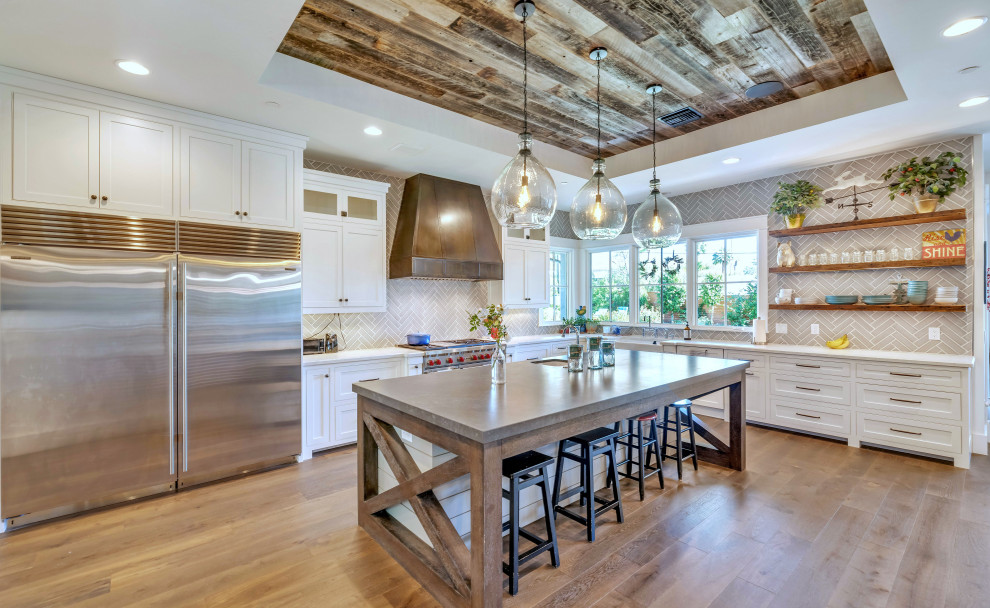 This is an example of a country l-shaped kitchen in Phoenix with a farmhouse sink, shaker cabinets, white cabinets, grey splashback, subway tile splashback, stainless steel appliances, medium hardwood floors, with island, brown floor, white benchtop and recessed.