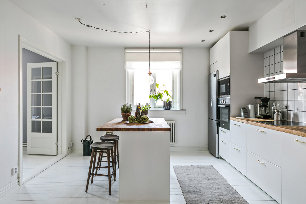Scandinavian galley separate kitchen in Gothenburg with flat-panel cabinets, white cabinets, wood benchtops, white splashback, stainless steel appliances, painted wood floors, with island, white floor and brown benchtop.