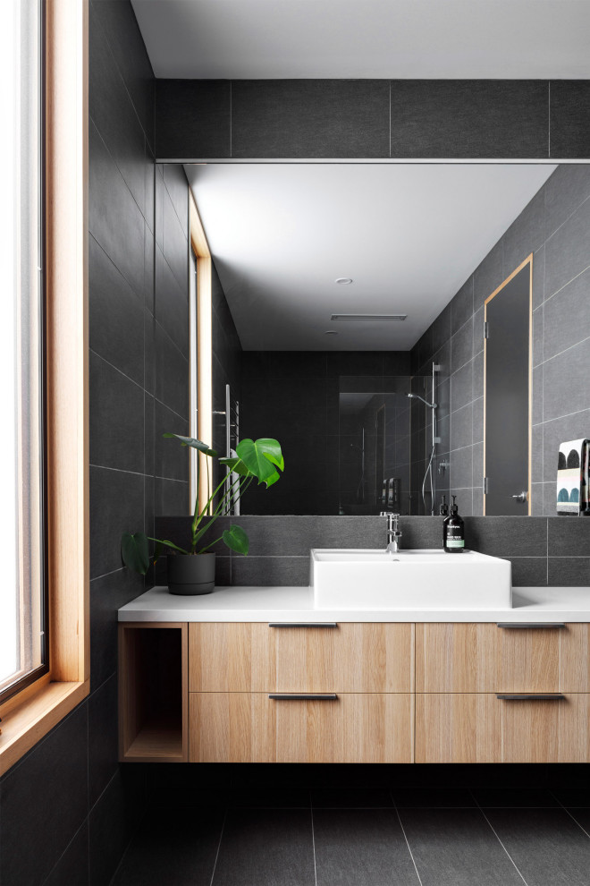 Идея дизайна: ванная комната среднего размера в современном стиле с плоскими фасадами, светлыми деревянными фасадами, накладной ванной, открытым душем, унитазом-моноблоком, черной плиткой, керамической плиткой, черными стенами, полом из керамической плитки, накладной раковиной, столешницей из искусственного камня, черным полом, открытым душем, белой столешницей, тумбой под одну раковину и встроенной тумбой