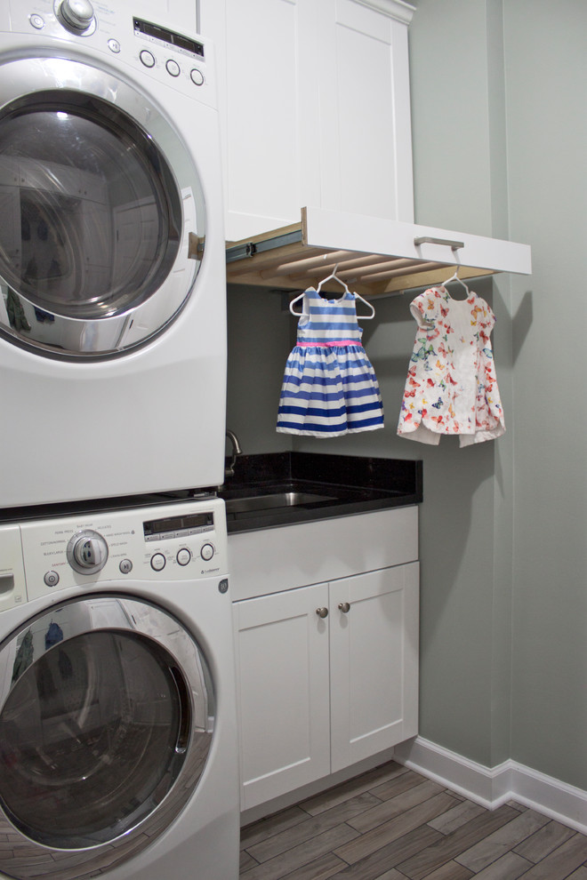 Photo of a mid-sized transitional utility room in Chicago with an undermount sink, shaker cabinets, white cabinets, granite benchtops, grey walls, ceramic floors, a stacked washer and dryer and grey floor.