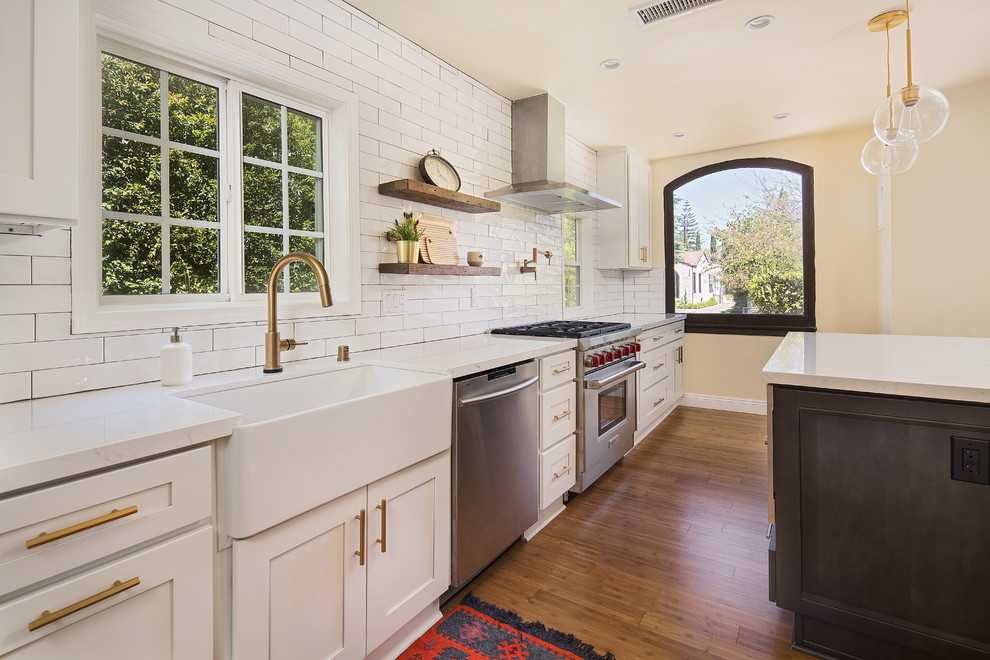 This is an example of a mid-sized country single-wall open plan kitchen in Los Angeles with a farmhouse sink, shaker cabinets, white cabinets, quartz benchtops, white splashback, porcelain splashback, stainless steel appliances, light hardwood floors, with island, brown floor and yellow benchtop.