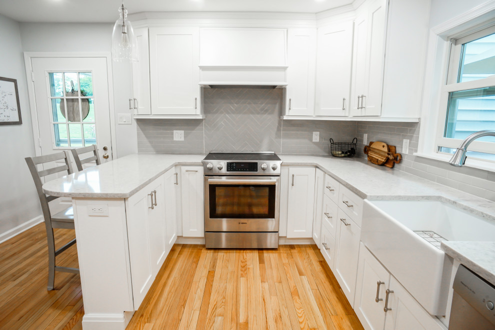 Photo of a traditional kitchen/diner in Philadelphia with a belfast sink, shaker cabinets, white cabinets, engineered stone countertops, grey splashback, light hardwood flooring, a breakfast bar, orange floors and grey worktops.