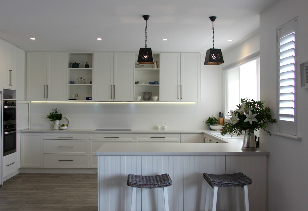 Photo of a mid-sized contemporary u-shaped open plan kitchen in Adelaide with shaker cabinets, white cabinets, solid surface benchtops, white splashback, stainless steel appliances, light hardwood floors and a peninsula.