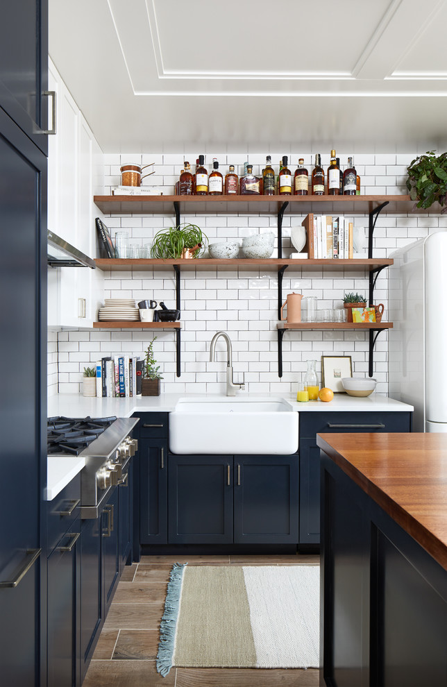 Small eclectic l-shaped open plan kitchen in DC Metro with a farmhouse sink, shaker cabinets, blue cabinets, quartz benchtops, white splashback, ceramic splashback, white appliances, porcelain floors, with island and brown floor.
