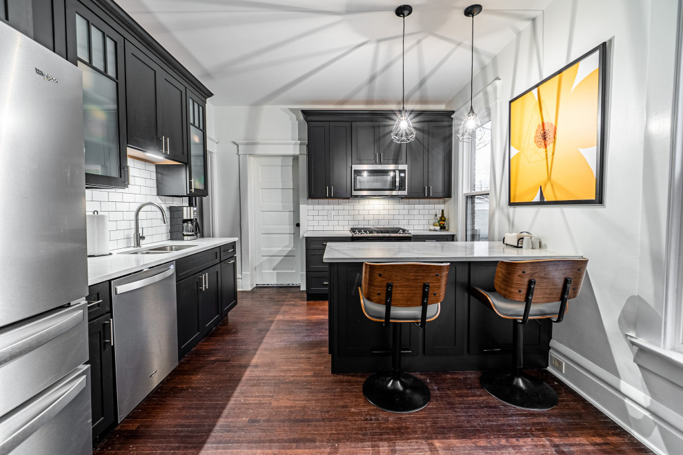 Photo of a mid-sized transitional l-shaped kitchen in Columbus with a double-bowl sink, shaker cabinets, black cabinets, white splashback, subway tile splashback, stainless steel appliances, medium hardwood floors, a peninsula, brown floor and grey benchtop.