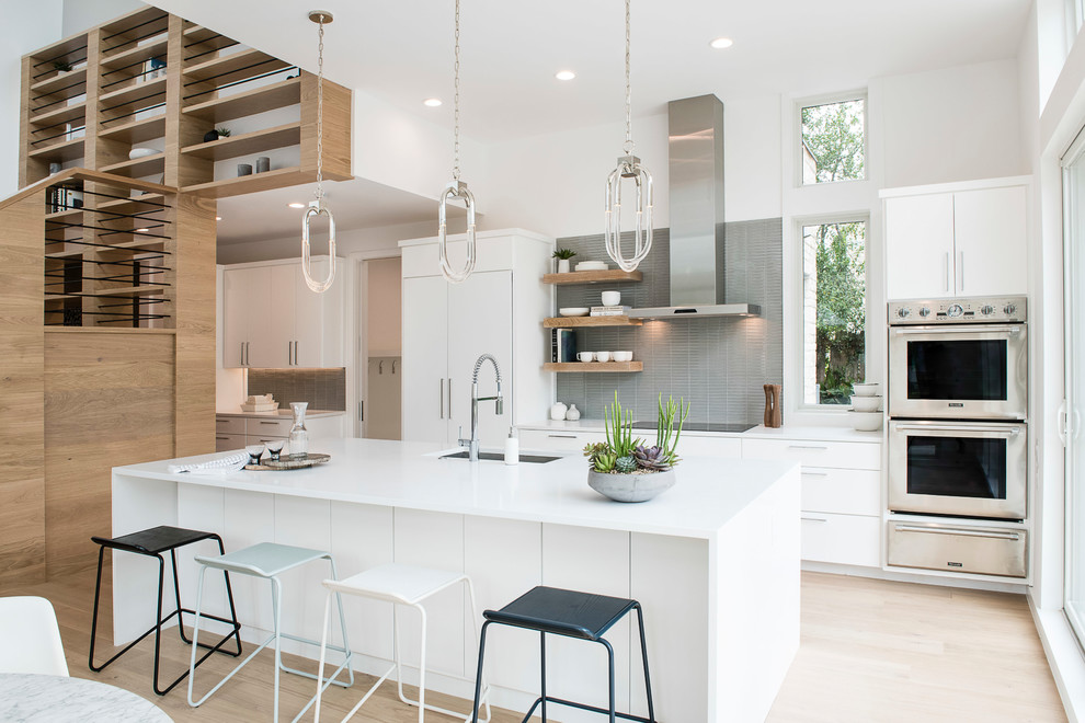 Photo of a mid-sized contemporary eat-in kitchen in Austin with an undermount sink, flat-panel cabinets, white cabinets, solid surface benchtops, grey splashback, glass tile splashback, stainless steel appliances, with island, white benchtop and light hardwood floors.