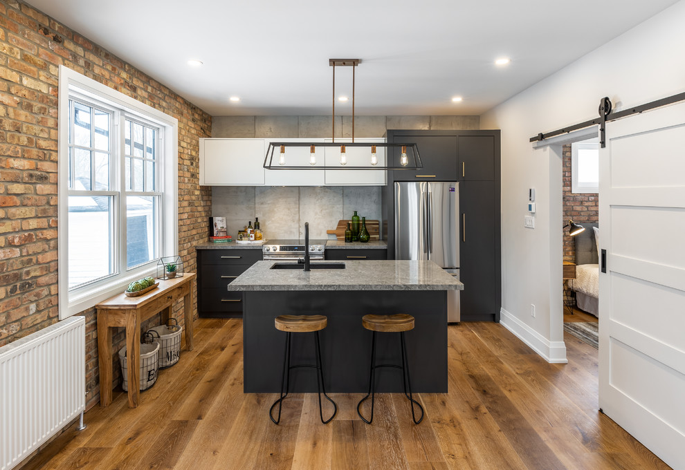 Photo of a small industrial galley kitchen in Toronto with an undermount sink, flat-panel cabinets, quartz benchtops, grey splashback, stainless steel appliances, medium hardwood floors, with island, brown floor, grey benchtop and black cabinets.