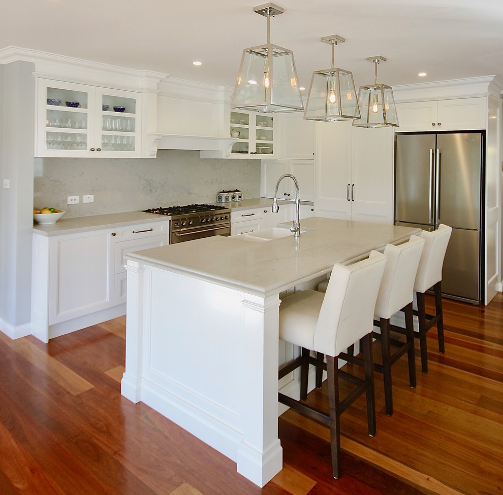 Photo of a large transitional l-shaped eat-in kitchen in Sydney with a farmhouse sink, recessed-panel cabinets, white cabinets, quartz benchtops, grey splashback, stone slab splashback, stainless steel appliances, medium hardwood floors, with island and brown floor.