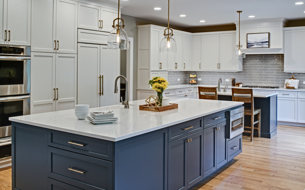 Photo of a large transitional u-shaped kitchen in Chicago with a farmhouse sink, recessed-panel cabinets, white cabinets, quartz benchtops, grey splashback, porcelain splashback, stainless steel appliances, light hardwood floors, multiple islands, brown floor and white benchtop.
