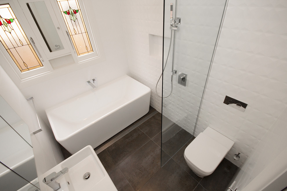 This is an example of a mid-sized modern master bathroom in Melbourne with a wall-mount sink, a freestanding tub, an open shower, a one-piece toilet, white tile, porcelain tile, white walls and porcelain floors.