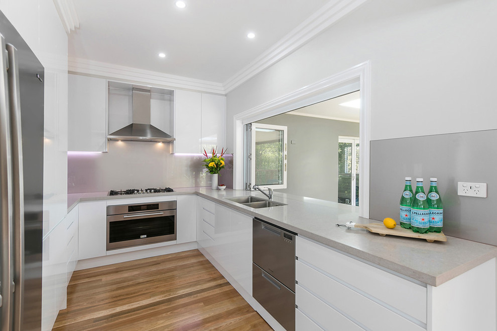 Photo of a mid-sized contemporary u-shaped kitchen in Sydney with an undermount sink, flat-panel cabinets, white cabinets, quartz benchtops, grey splashback, glass sheet splashback, stainless steel appliances, medium hardwood floors, no island and brown floor.