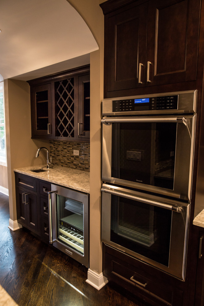 Home design - mid-sized transitional l-shaped dark wood floor home design idea in Chicago with an undermount sink, shaker cabinets, brown cabinets, granite countertops, gray backsplash, subway tile backsplash, stainless steel appliances and an island