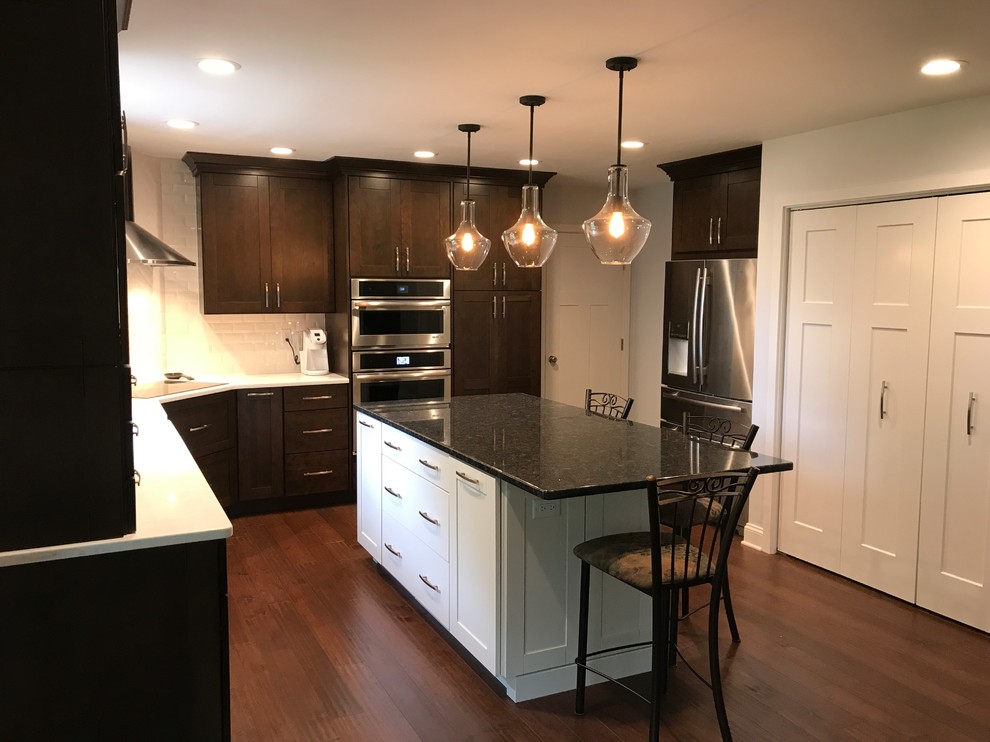 Mid-sized traditional l-shaped eat-in kitchen in Baltimore with a farmhouse sink, shaker cabinets, dark wood cabinets, quartz benchtops, white splashback, subway tile splashback, stainless steel appliances, dark hardwood floors, with island, brown floor and white benchtop.