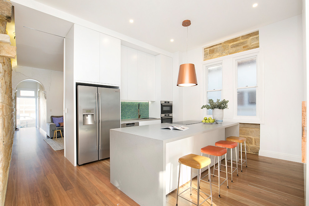 Contemporary single-wall kitchen in Sydney with white cabinets, green splashback, stainless steel appliances, medium hardwood floors, with island, brown floor, grey benchtop, an undermount sink, flat-panel cabinets, solid surface benchtops and mosaic tile splashback.