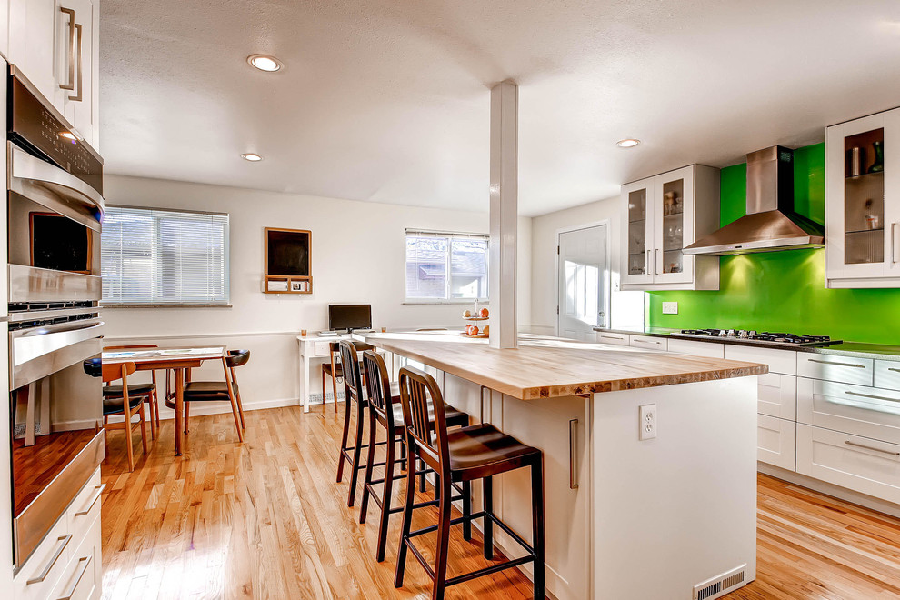 Photo of a mid-sized midcentury u-shaped eat-in kitchen in Denver with green splashback, stainless steel appliances, white cabinets, wood benchtops, a single-bowl sink, recessed-panel cabinets, glass sheet splashback, medium hardwood floors and with island.