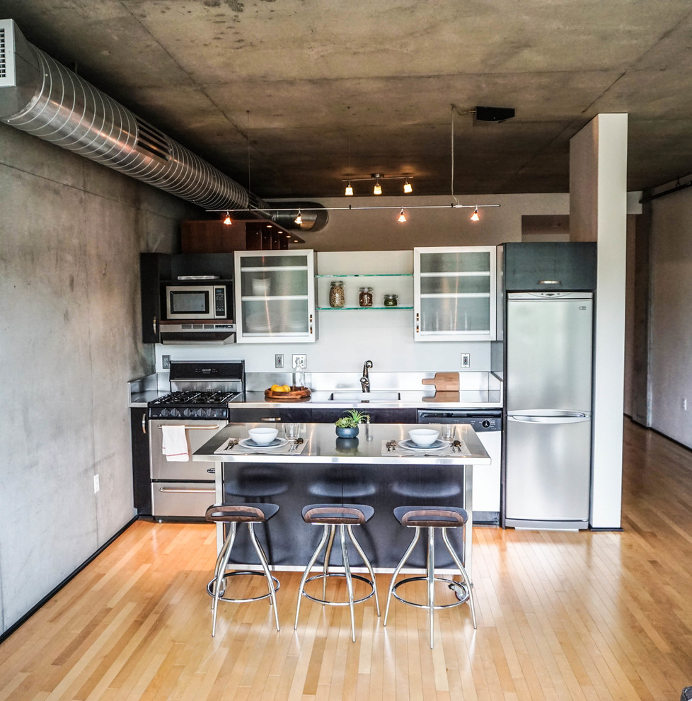 Design ideas for a mid-sized industrial single-wall open plan kitchen in Portland with a single-bowl sink, glass-front cabinets, stainless steel cabinets, stainless steel benchtops, stainless steel appliances, light hardwood floors and with island.