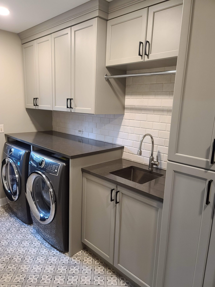 This is an example of a large transitional laundry room in Chicago with grey cabinets, quartz benchtops, marble splashback, porcelain floors, grey floor, grey benchtop and a side-by-side washer and dryer.