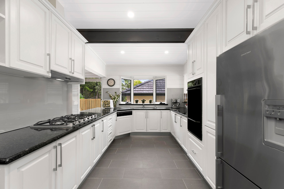 Transitional galley separate kitchen in Sydney with a drop-in sink, raised-panel cabinets, white cabinets, grey splashback, glass sheet splashback, stainless steel appliances, white walls, no island and grey floor.
